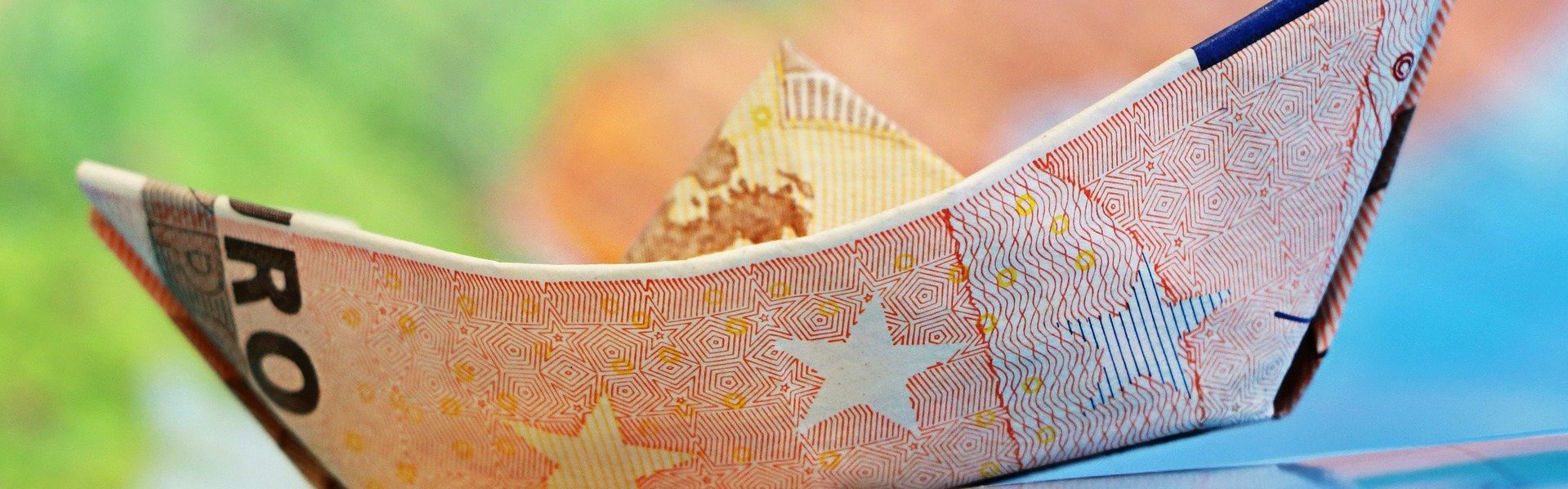 Das Bild zeigt ein aus einem Geldschein gebasteltes Schiff vor einem bunten Hintergrund.