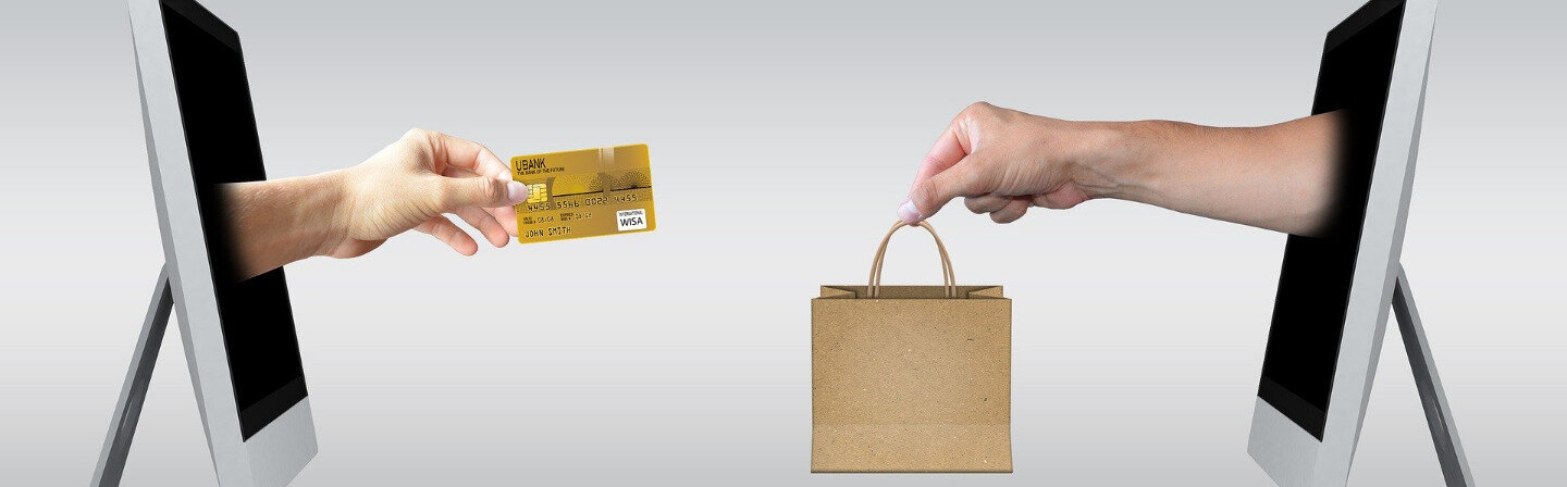 Das Bild zeigt zwei Arme, die jeweils aus einem Computer-Bildschirm herausragen. Die eine Hand hält eine Kreditkarte, die andere eine Einkaufstasche.